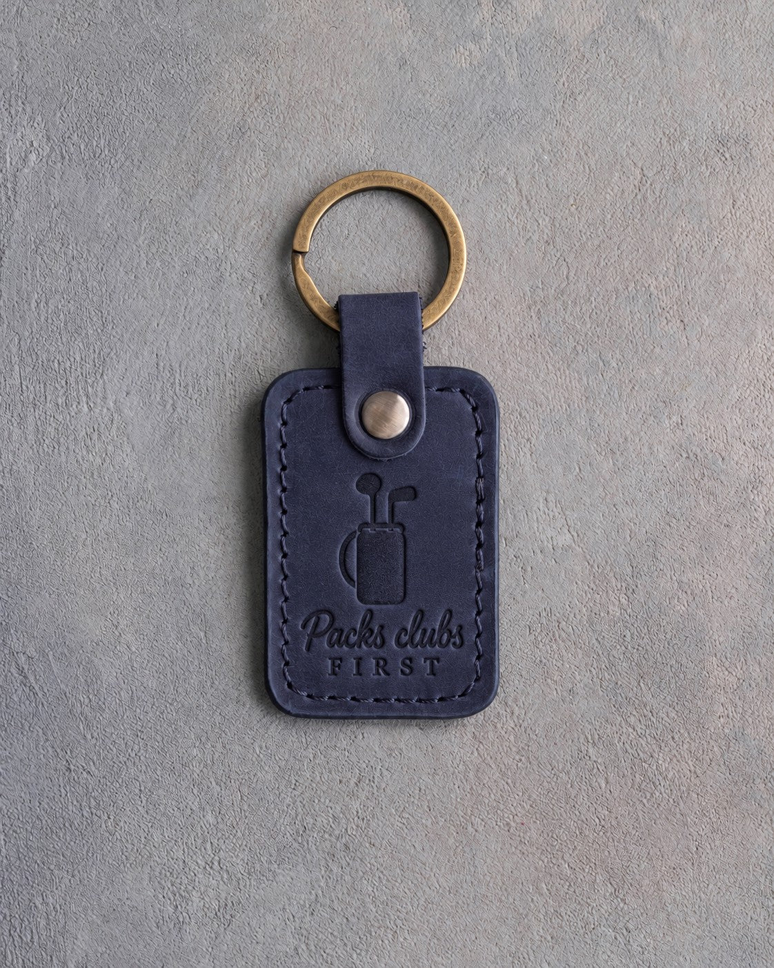 Blueberry navy leather keychain debossed with a golf bag holding two clubs above Packs clubs FIRST, on gray plaster