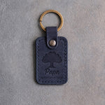 Blueberry navy leather keychain debossed with a solid oak tree above Papa and ROOTS RUN DEEP, on gray concrete