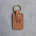 Caramel leather keychain debossed with a Papillon silhouette above THE PAP, on gray concrete
