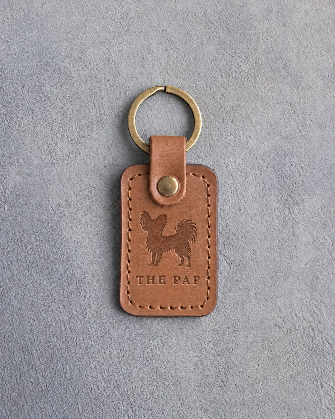Caramel leather keychain debossed with a Papillon silhouette above THE PAP, on gray concrete