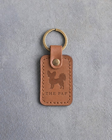 Caramel leather keychain debossed with a Papillon silhouette above THE PAP, on gray concrete