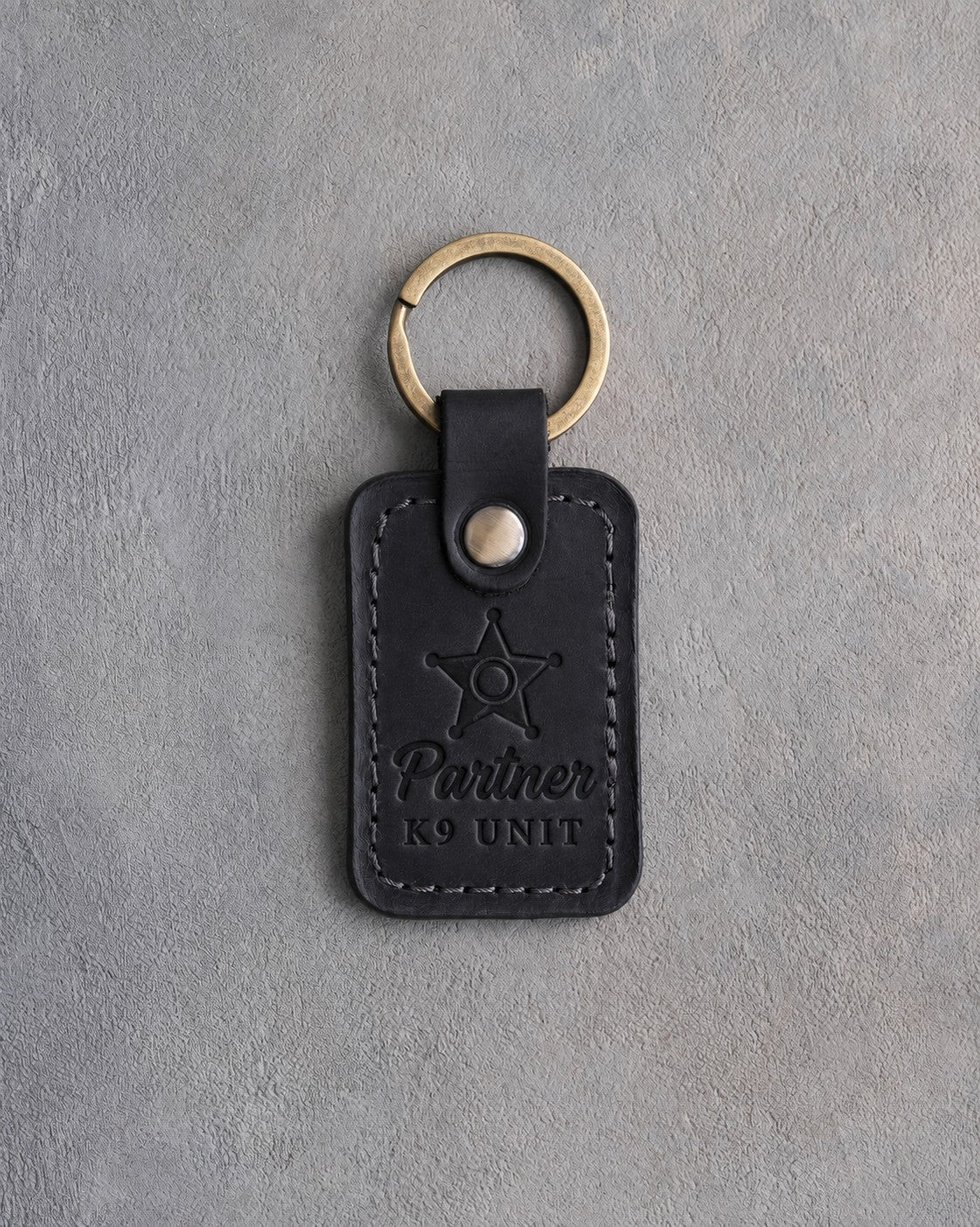 Black leather keychain blind debossed with a five-pointed star badge and the words Partner K9 UNIT, on gray concrete