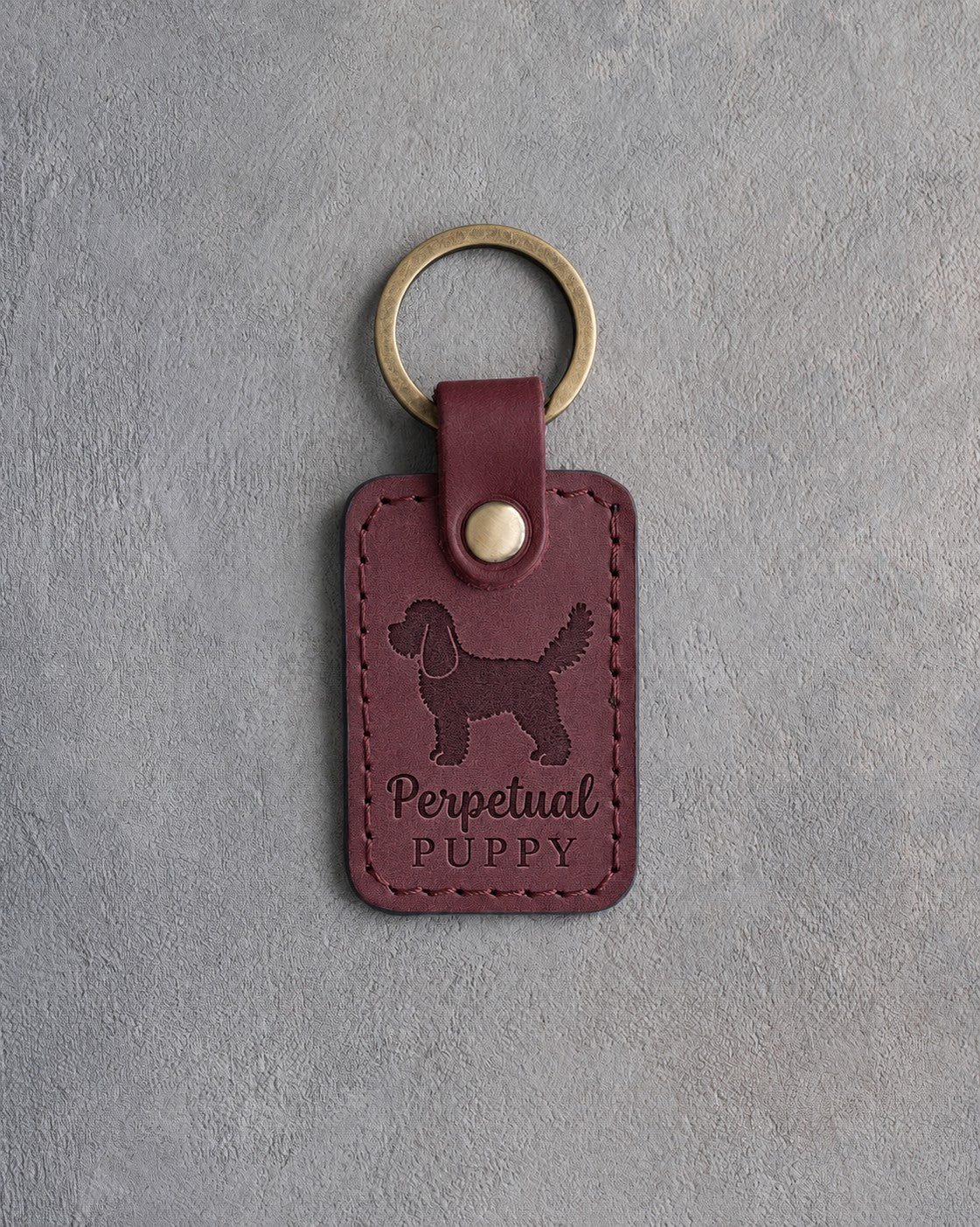 Sangria wine-red leather keychain debossed with a cockapoo and the words Perpetual PUPPY, on gray concrete