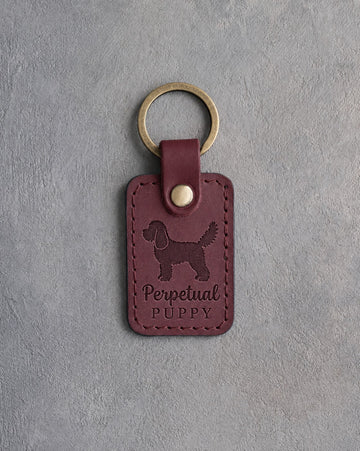 Sangria wine-red leather keychain debossed with a cockapoo and the words Perpetual PUPPY, on gray concrete