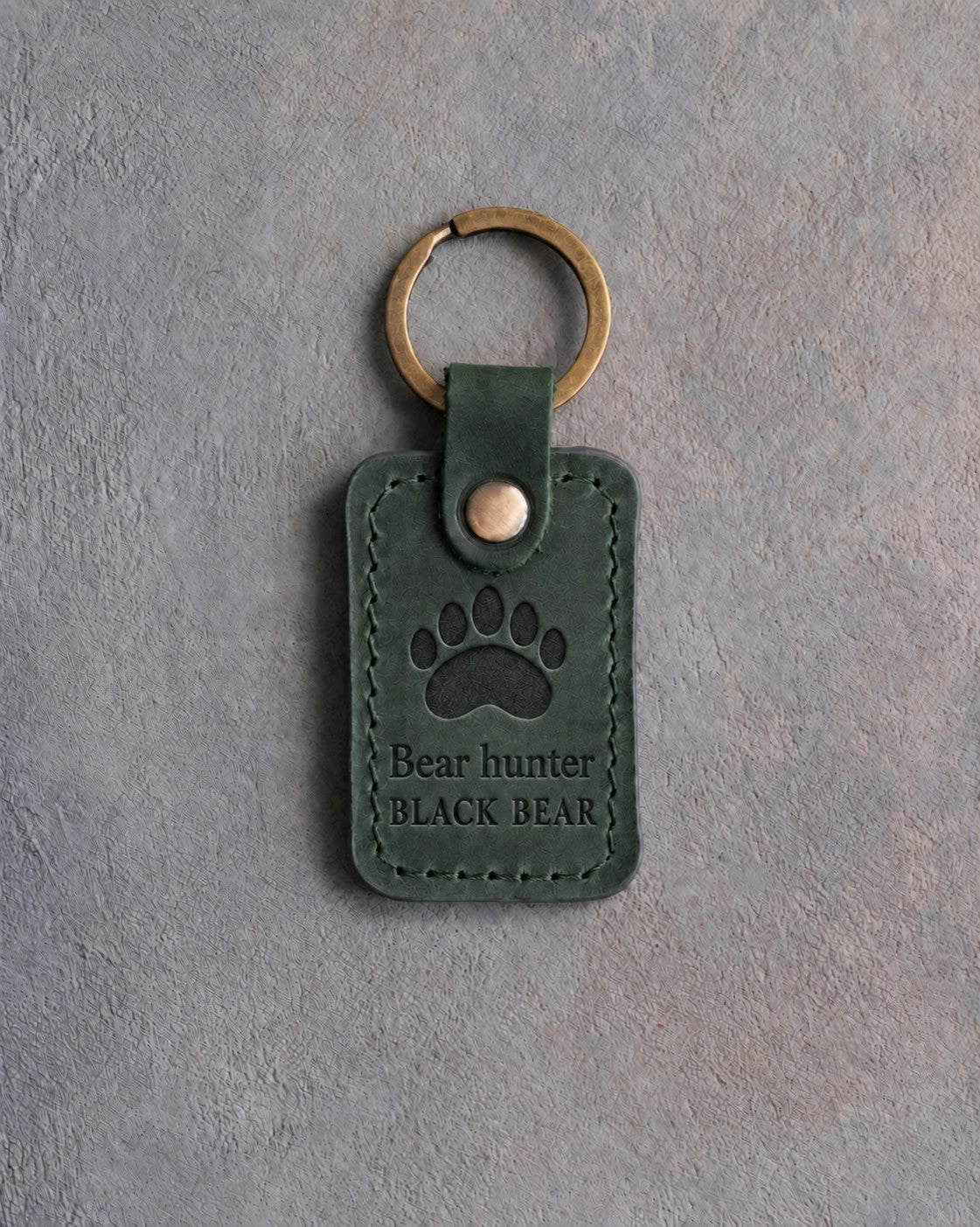 Forest green leather keychain debossed with a black bear paw track above the words Bear hunter and BLACK BEAR, on gray concrete