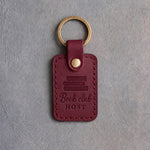 Sangria wine red leather keychain debossed with a stack of books above Book club and HOST, on gray concrete