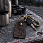 Levi BOWHUNTER keychain in dark brown leather hanging from the grip of a compound bow against a tree trunk