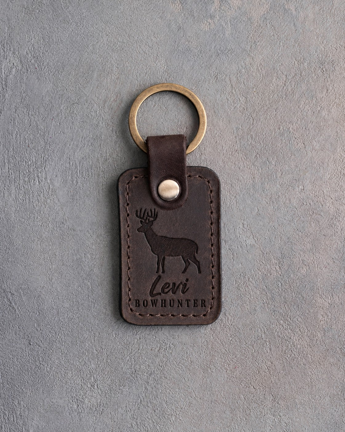 Dark espresso leather keychain debossed with a whitetail buck above the name Levi and BOWHUNTER, on gray plaster