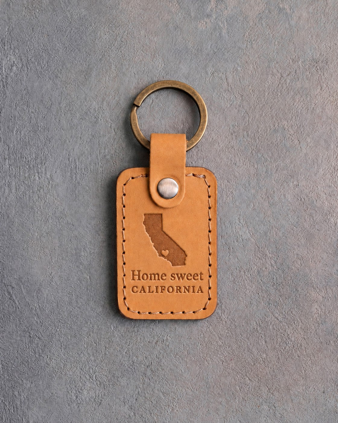 Indian Summer tan leather keychain debossed with a California silhouette and heart above the words Home sweet and CALIFORNIA, on gray concrete