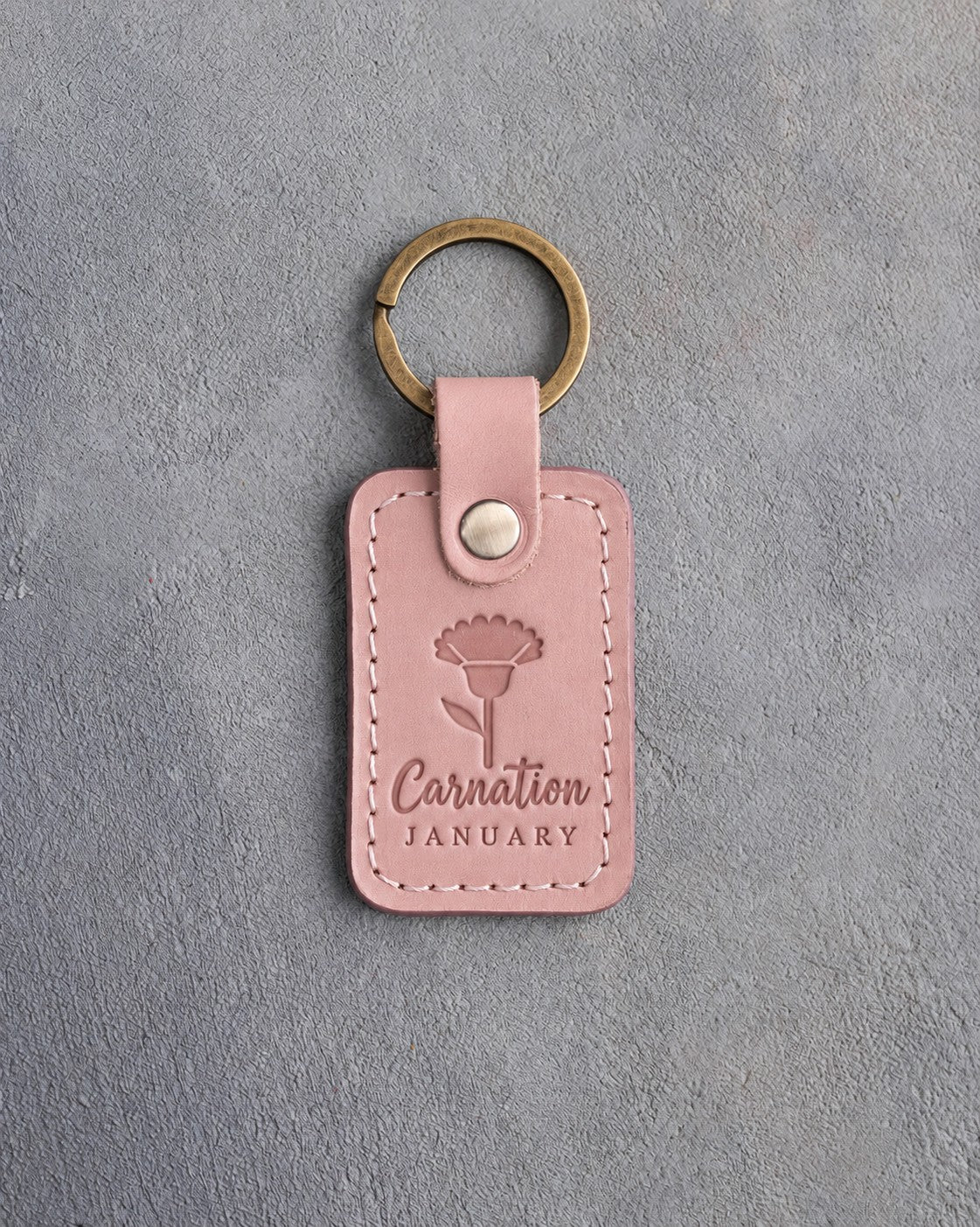Blush pink leather keychain debossed with a carnation above Carnation and JANUARY, on gray concrete