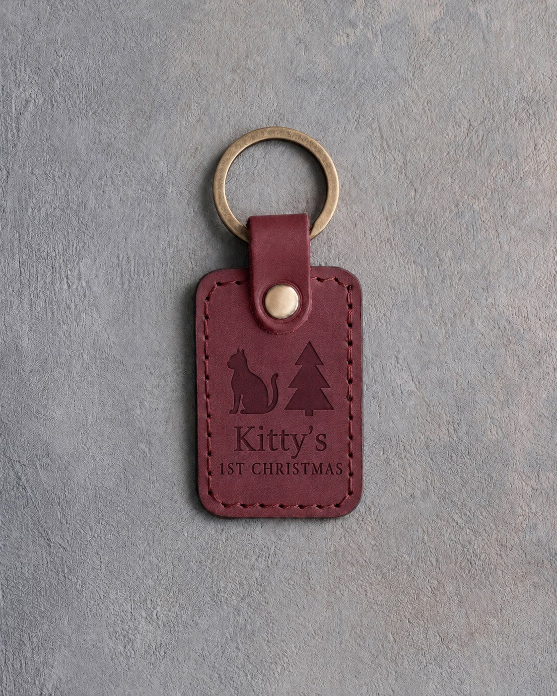Sangria red leather keychain debossed with a small cat beside a Christmas tree above Kitty's and 1ST CHRISTMAS, on gray concrete
