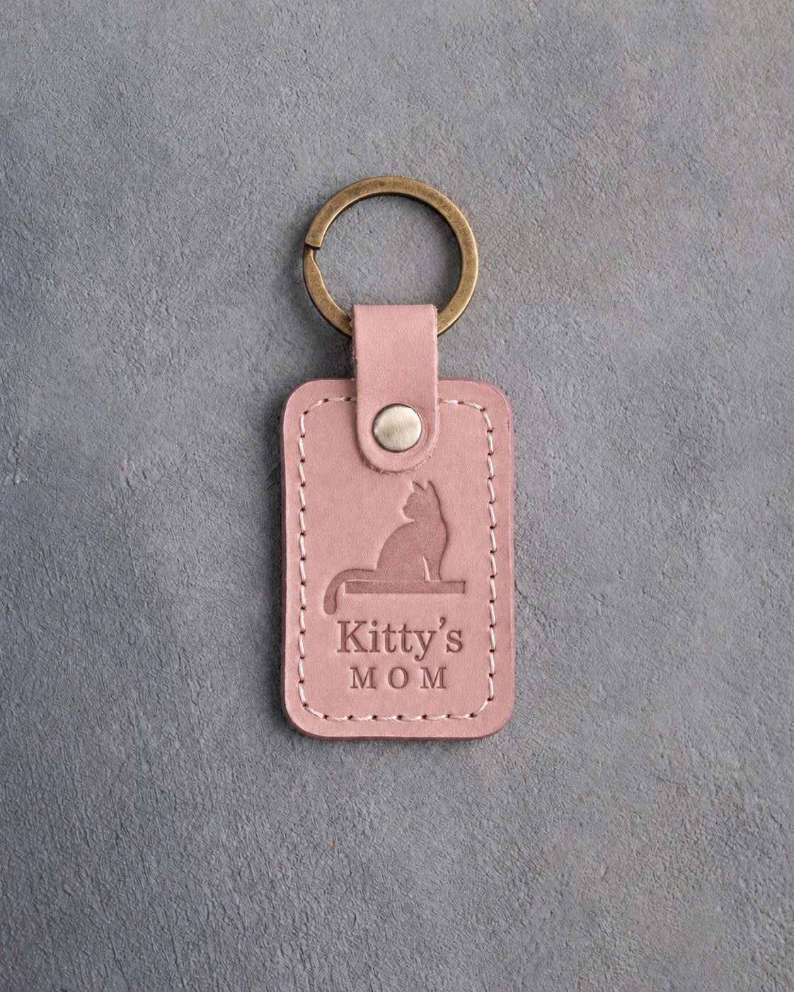 Blush pink leather keychain debossed with a cat on a windowsill above Kitty's and MOM, on gray concrete