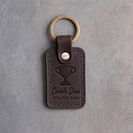 Dark espresso leather keychain debossed with a trophy cup above Coach Dan and SPRING 2026, on gray plaster