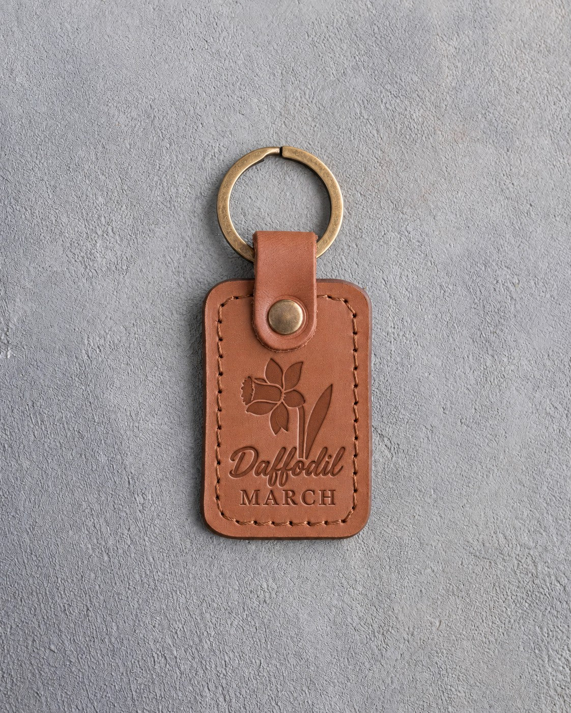 Caramel leather keychain debossed with a daffodil above Daffodil and MARCH, on gray concrete
