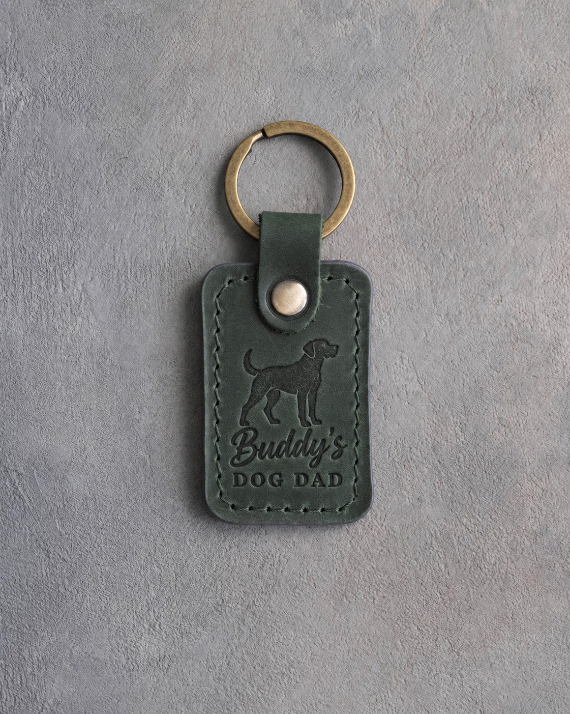Forest green leather keychain debossed with an alert standing dog above Buddy's and DOG DAD, on gray concrete
