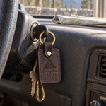 Dark brown Driving Home keychain hanging from a car ignition with a snowy highway at dusk through the windshield