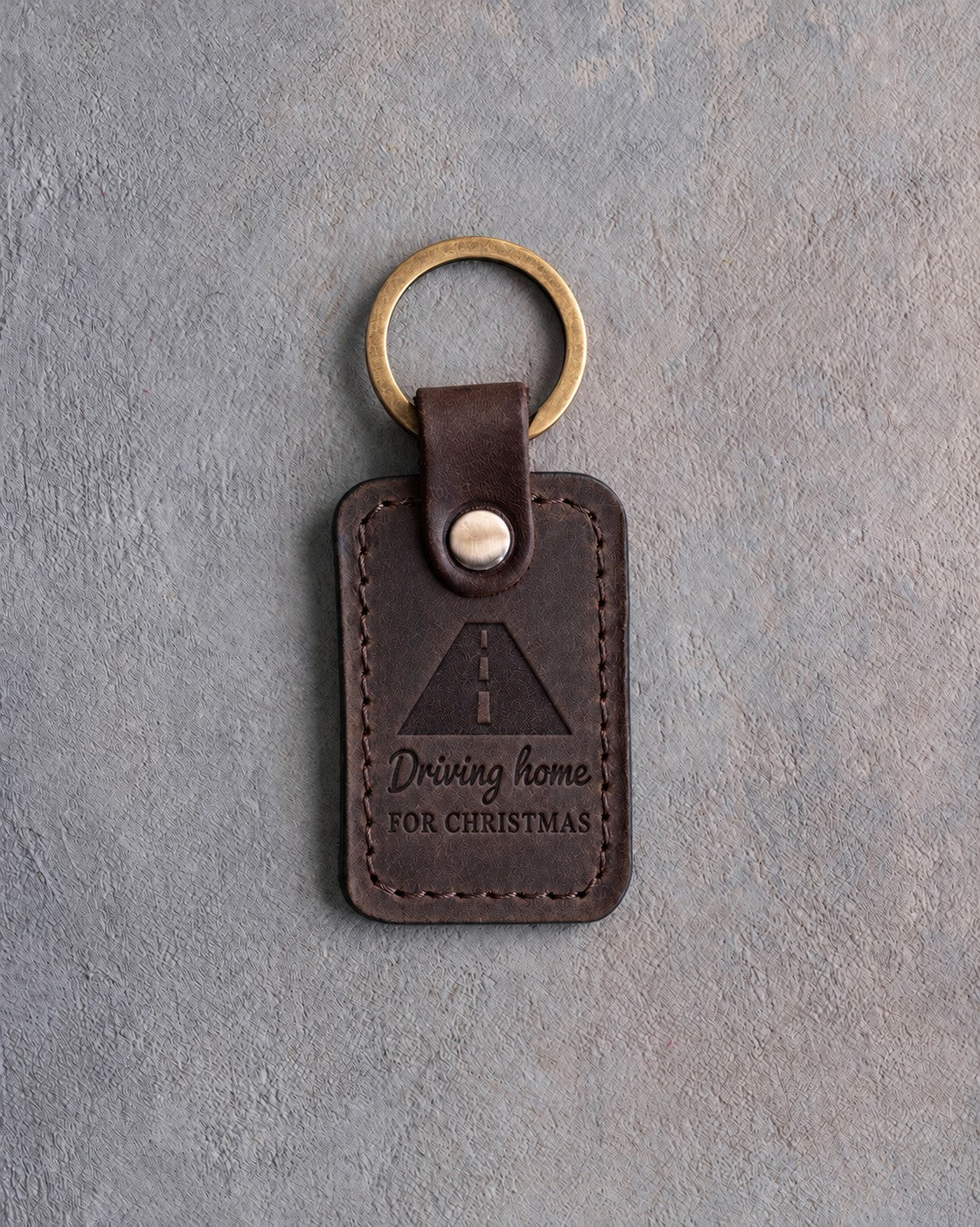 Dark espresso leather keychain debossed with an open road in perspective above the words Driving home and FOR CHRISTMAS, on gray concrete