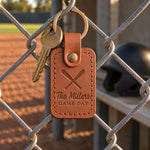 Caramel The Millers GAME DAY keychain hanging on a chain-link dugout fence above a raked infield
