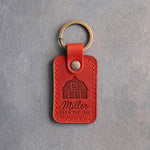 Poppy red leather keychain debossed with a gambrel barn above the name Miller and FARM EST 1946, on gray concrete