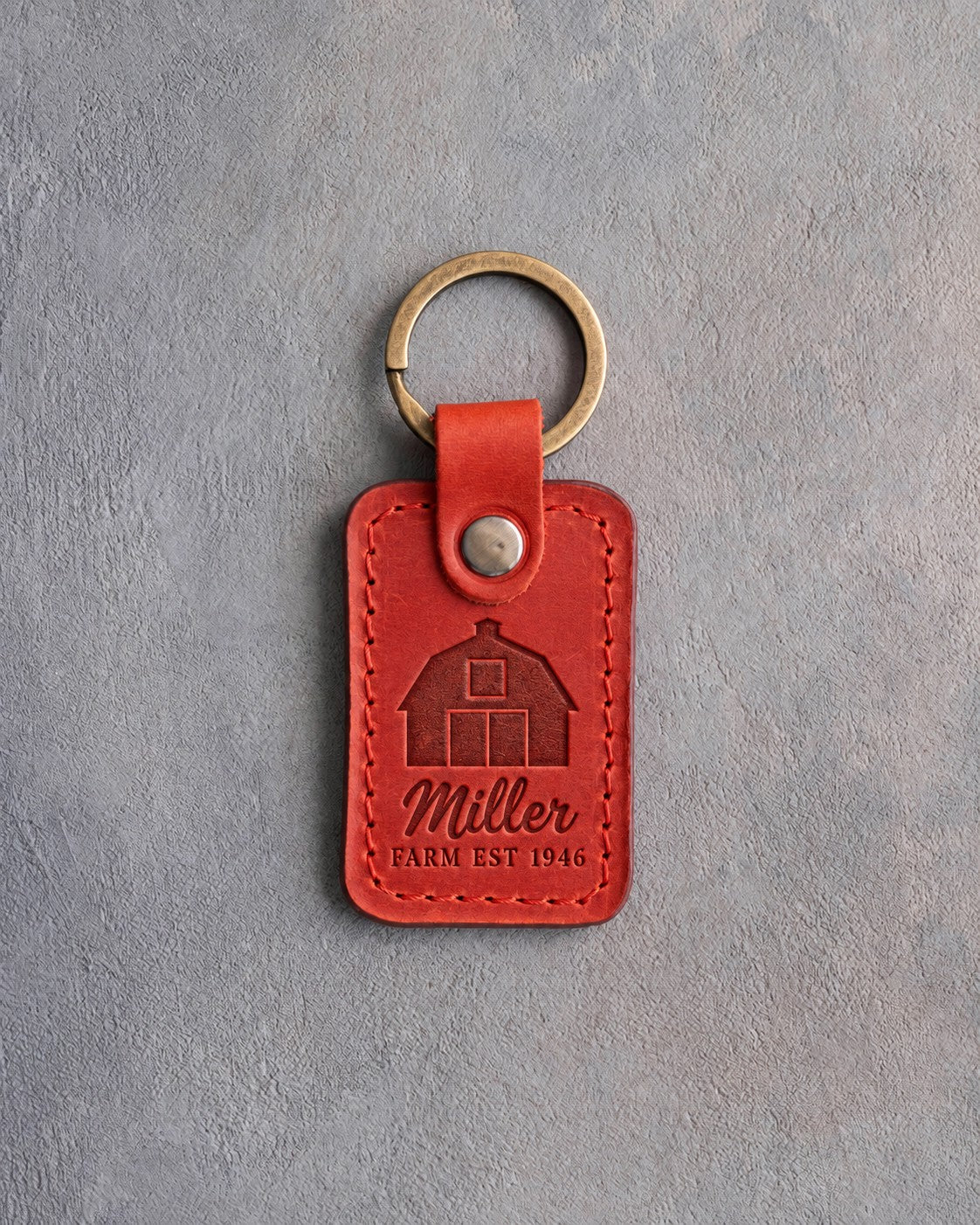 Poppy red leather keychain debossed with a gambrel barn above the name Miller and FARM EST 1946, on gray concrete