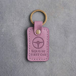 Lilac leather keychain debossed with a steering wheel above the words Keys to my and FIRST CAR, on gray concrete