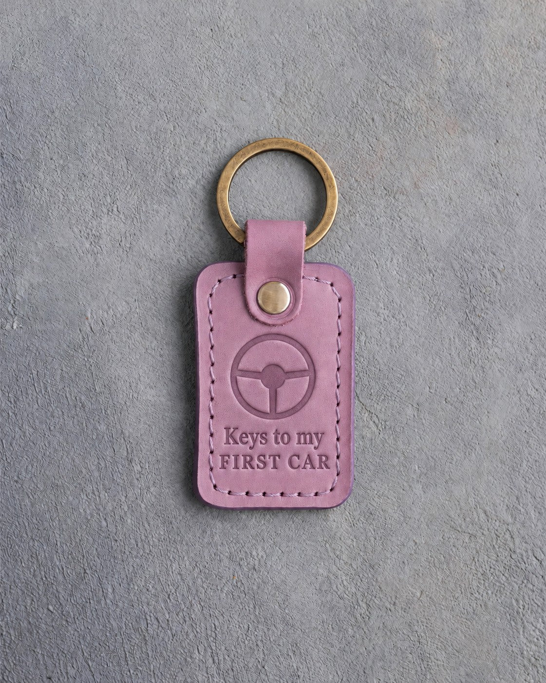 Lilac leather keychain debossed with a steering wheel above the words Keys to my and FIRST CAR, on gray concrete