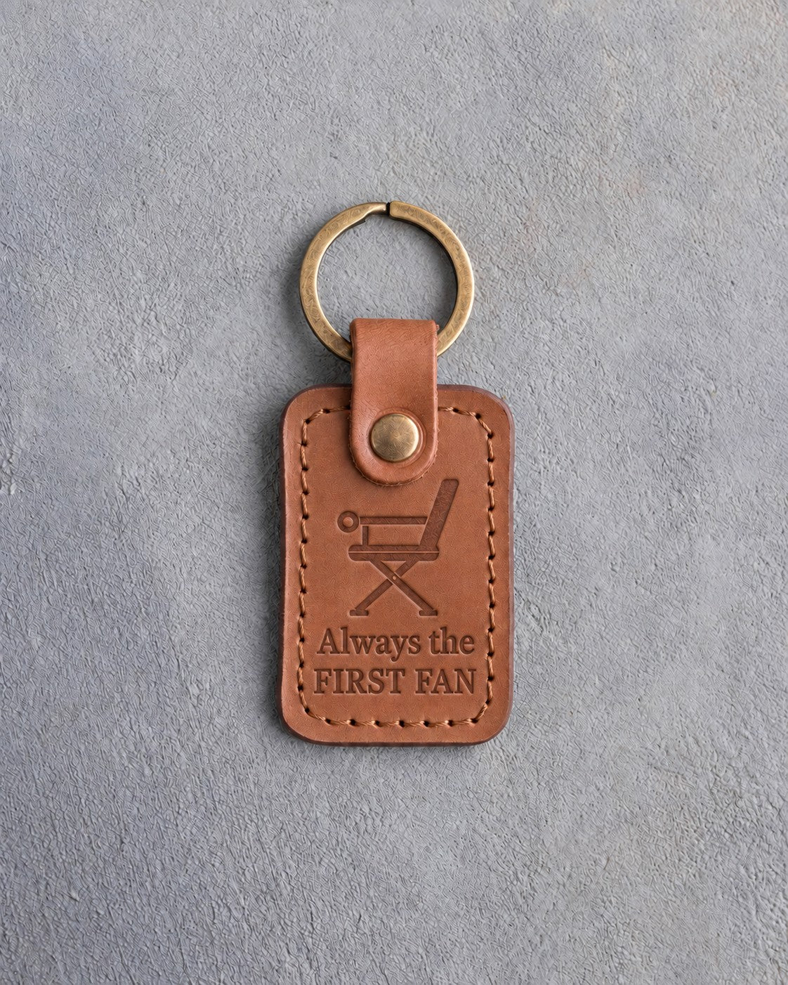 Caramel leather keychain debossed with a folding sideline chair above Always the and FIRST FAN, on gray concrete