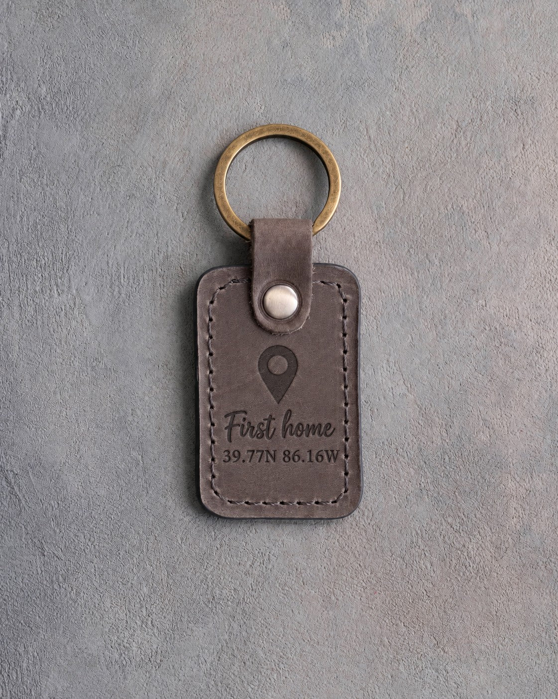 Country gray leather keychain debossed with a map pin above First home and 39.77N 86.16W, on gray concrete
