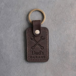 Dark espresso leather keychain debossed with crossed wrenches above the words Dad's and GARAGE, on gray concrete