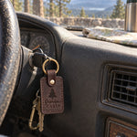 Dark brown Grandpa's Cabin keychain hanging from a nail by a log cabin door with a lake behind