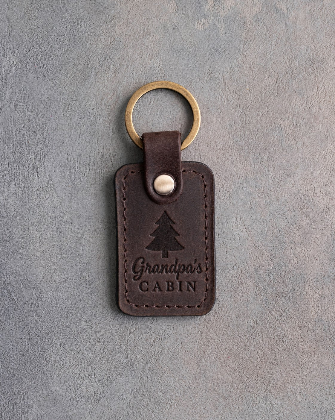 Dark espresso leather keychain debossed with a pine tree above Grandpa's and CABIN, on gray concrete