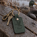 Reid GREENHEAD keychain in forest green leather on a blind bag zipper beside a wooden duck call