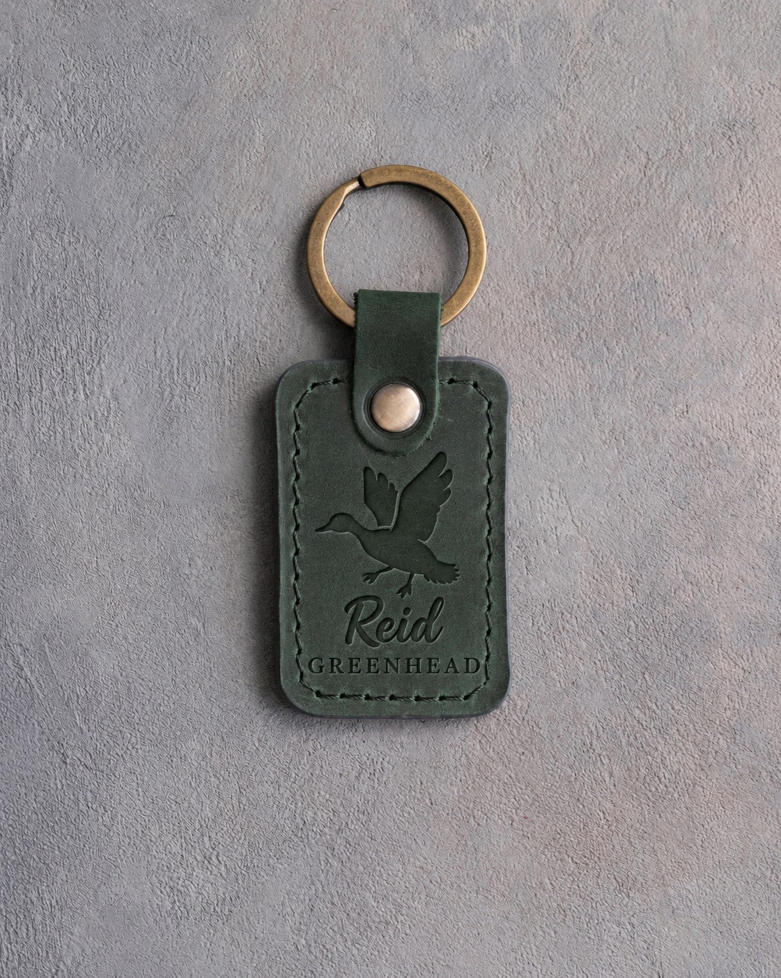 Forest green leather keychain debossed with a mallard cupped to land above the name Reid and GREENHEAD, on gray plaster