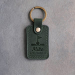 Forest green leather keychain debossed with a golf ball in the cup under a pin flag above the name Mike and ACE · HOLE 12, on gray plaster