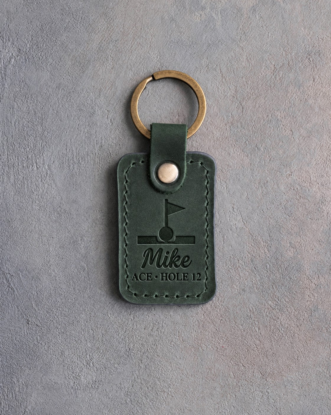 Forest green leather keychain debossed with a golf ball in the cup under a pin flag above the name Mike and ACE · HOLE 12, on gray plaster