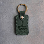 Forest green leather keychain debossed with a golf ball in the cup under a pin flag above the words Hole in one and CLUB, on gray concrete