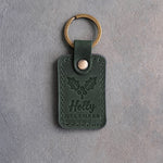 Forest green leather keychain debossed with a holly sprig above Holly and DECEMBER, on gray concrete