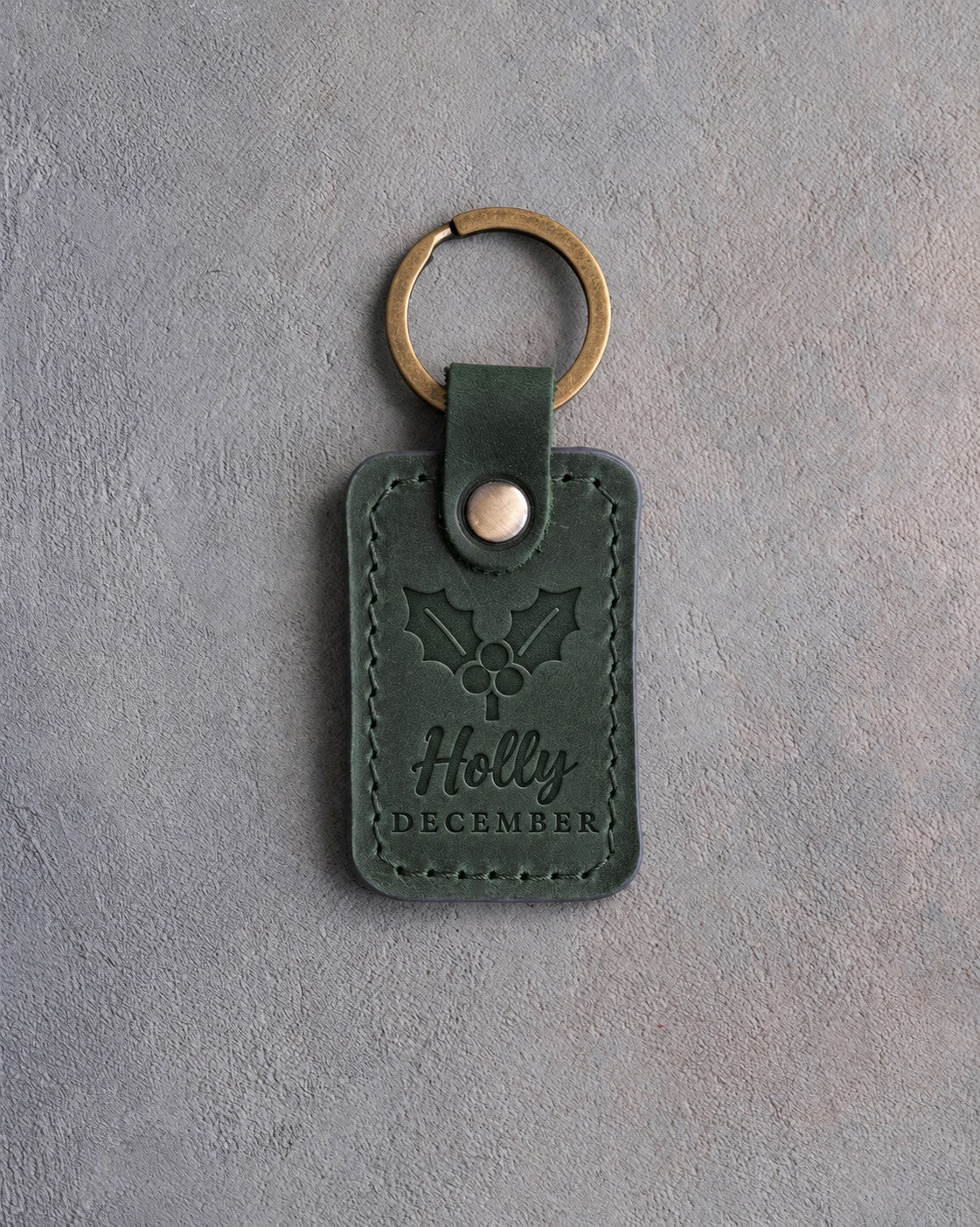 Forest green leather keychain debossed with a holly sprig above Holly and DECEMBER, on gray concrete