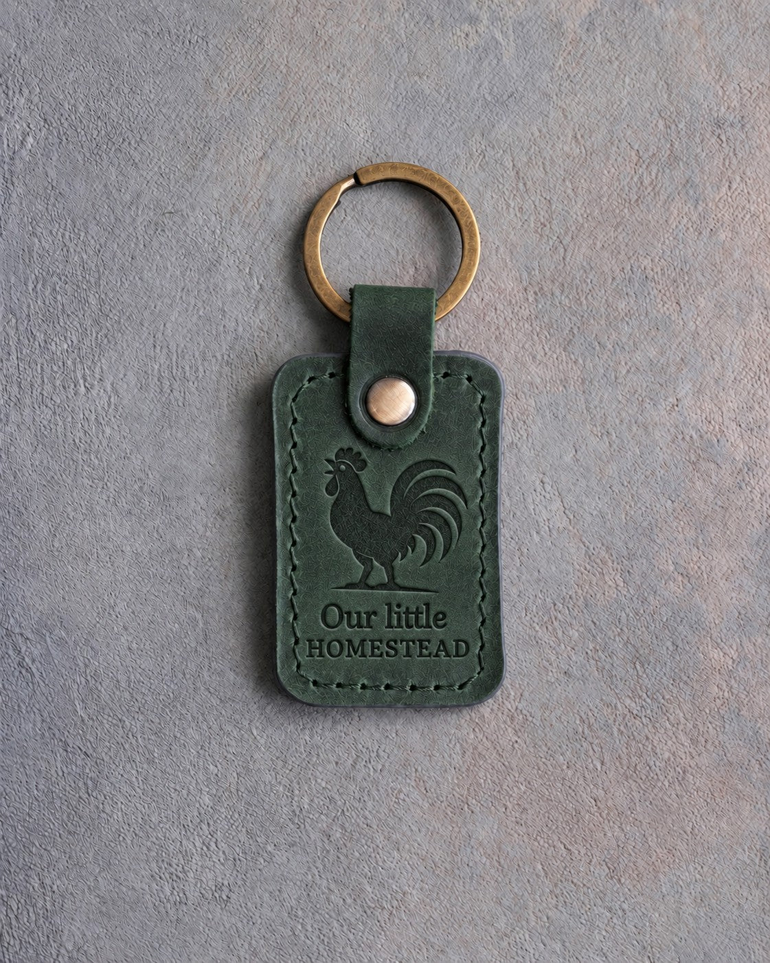 Forest green leather keychain debossed with a crowing rooster above the words Our little and HOMESTEAD, on gray concrete