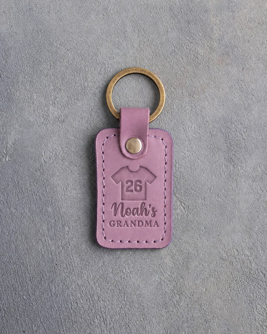 Lilac leather keychain debossed with a jersey numbered 26 above the name Noah's and GRANDMA, on gray plaster