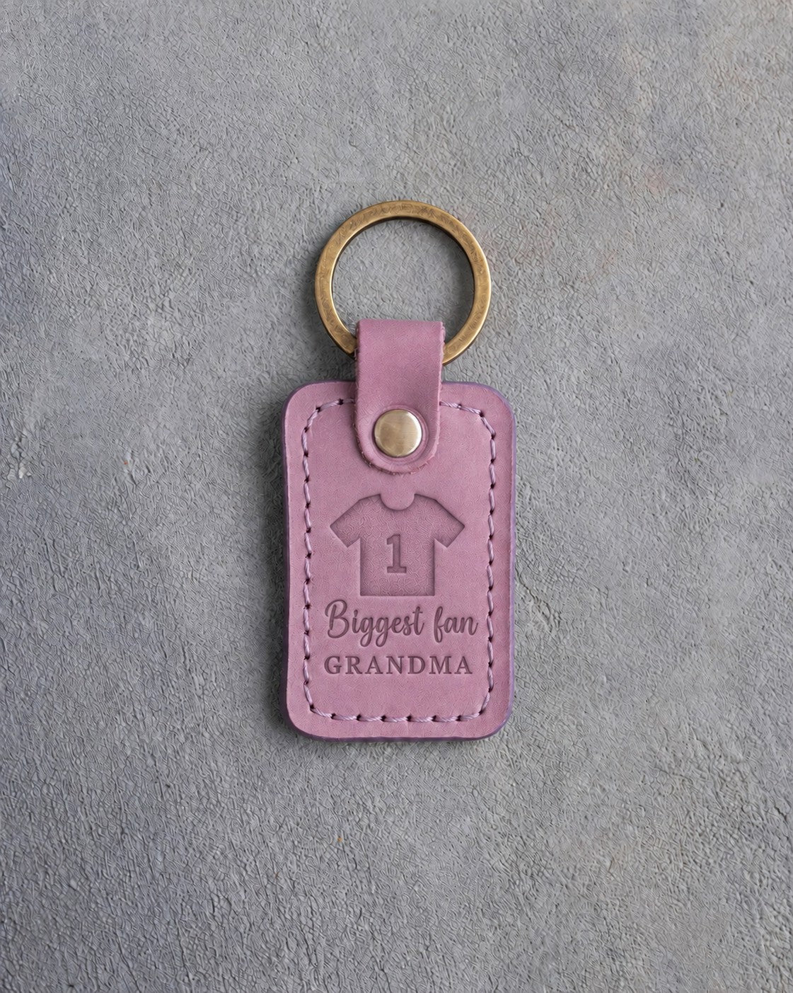 Lilac leather keychain debossed with a jersey numbered 1 above the words Biggest fan and GRANDMA, on gray concrete