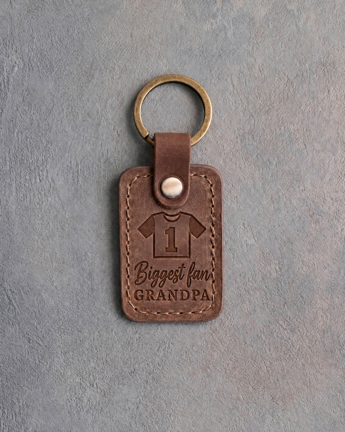 Sicilian brown leather keychain debossed with a jersey numbered 1 above the words Biggest fan and GRANDPA, on gray concrete