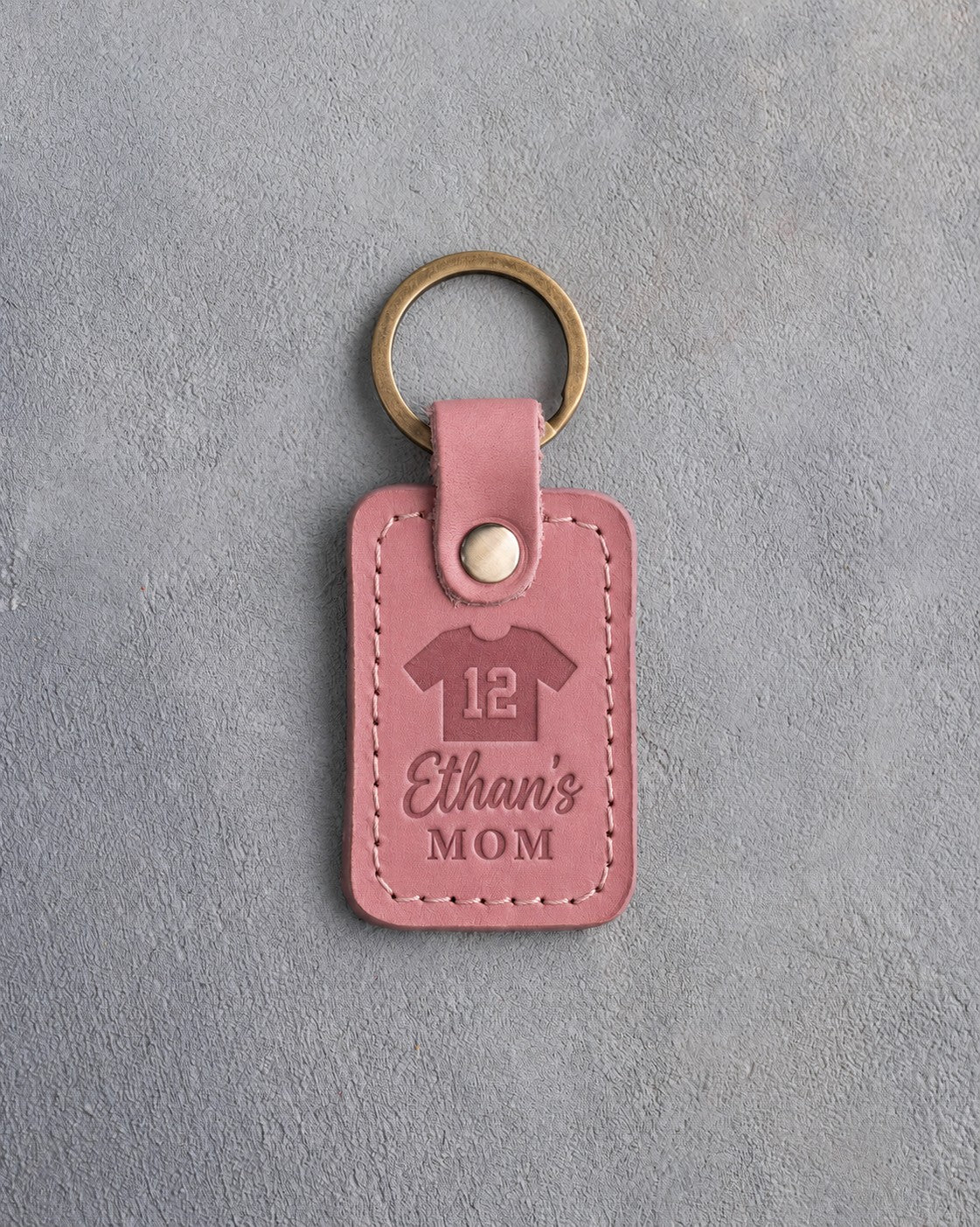 Pink Parisian Blossom leather keychain debossed with a jersey numbered 12 above the name Ethan's and MOM, on gray plaster