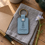 Turquoise Larkspur July keychain on a garden bench beside a jug of tall blue larkspur
