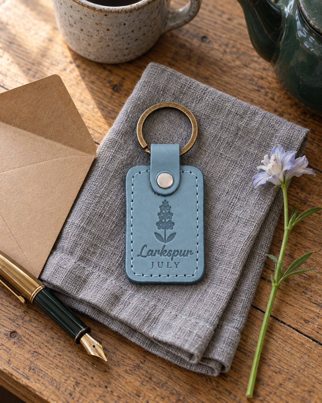 Turquoise Larkspur July keychain on a garden bench beside a jug of tall blue larkspur