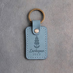 Turquoise leather keychain debossed with a larkspur spike above Larkspur and JULY, on gray concrete