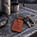 Hank LONGBEARD keychain in tan leather resting on a turkey vest beside a box call on an oak stump
