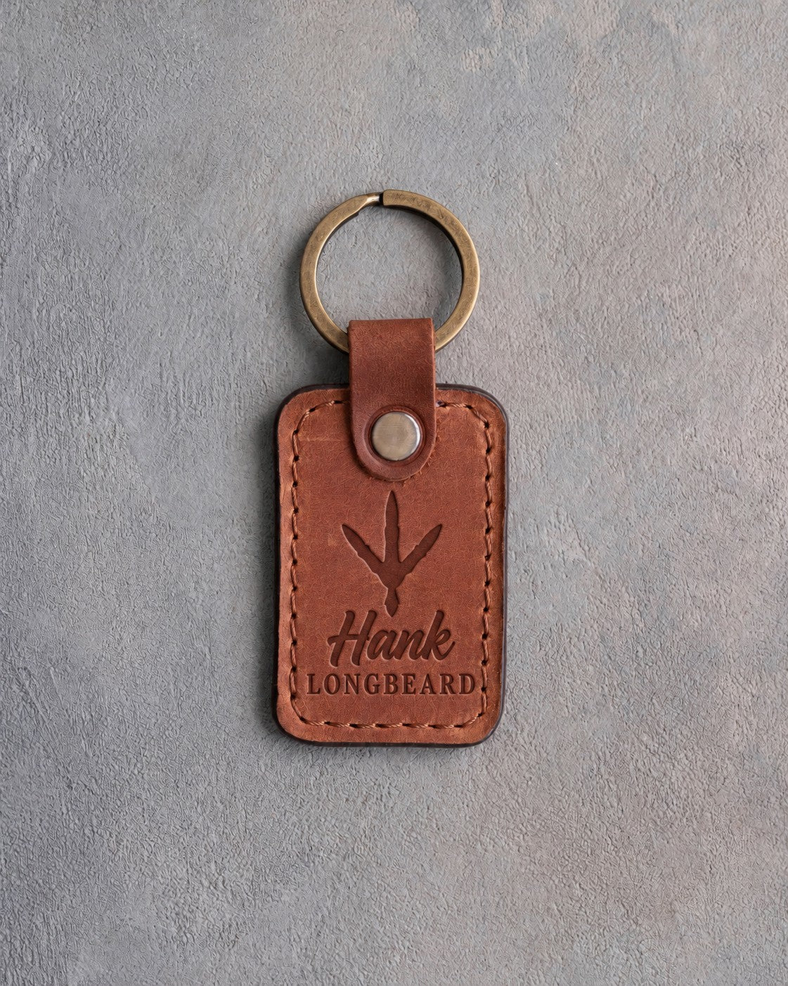 Warm tan leather keychain debossed with a wild turkey track above the name Hank and LONGBEARD, on gray plaster