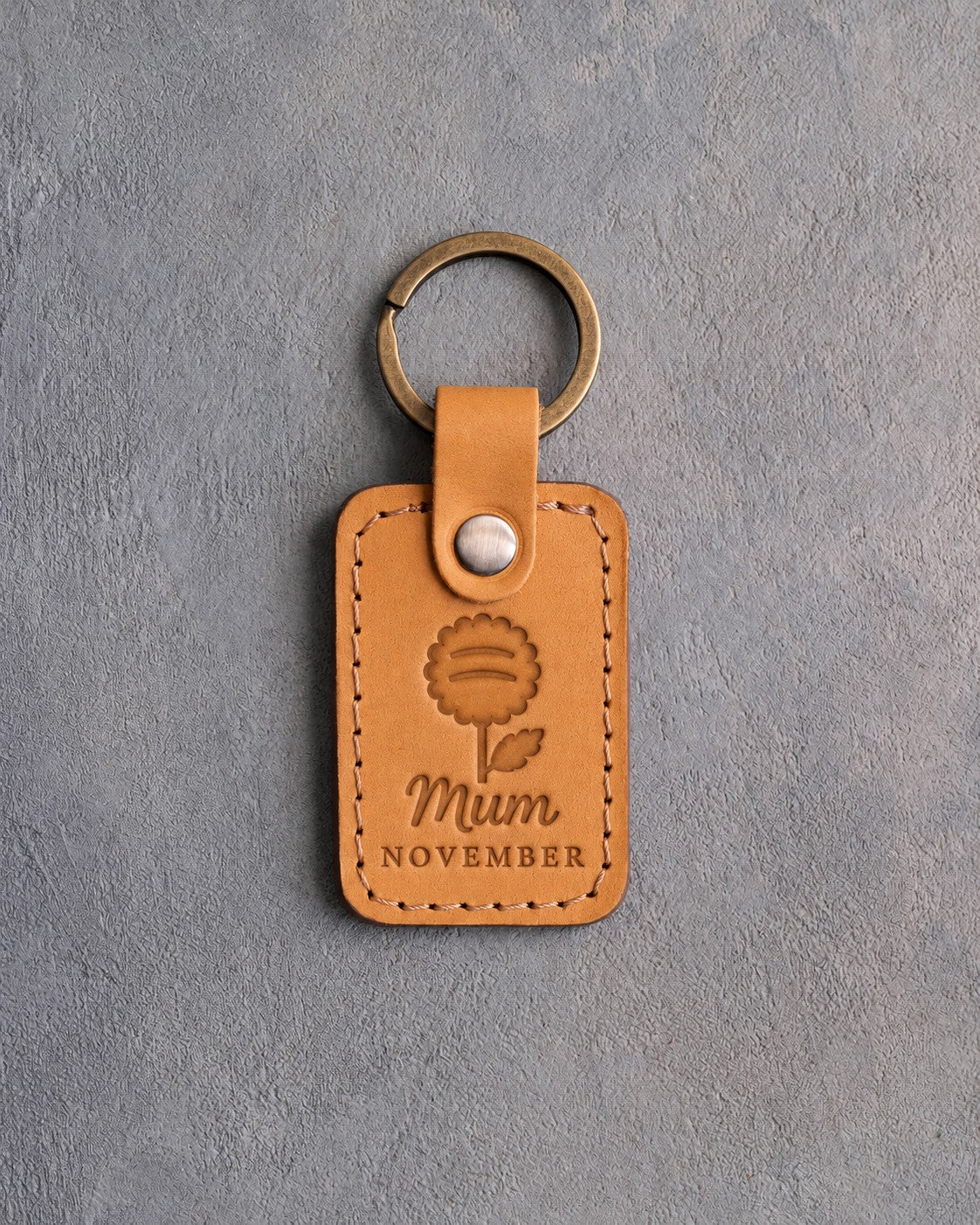 Indian Summer tan leather keychain debossed with a chrysanthemum above Mum and NOVEMBER, on gray concrete