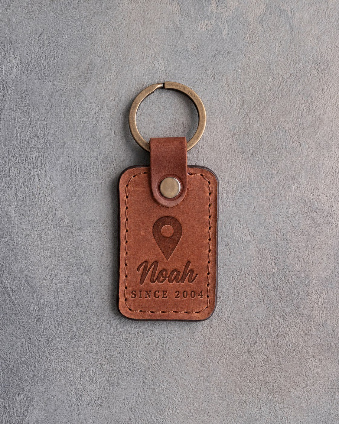 Sandy tan leather keychain debossed with a map pin above the name Noah and SINCE 2004, on gray concrete